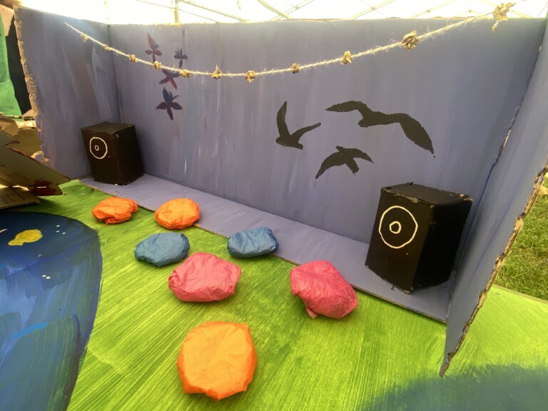 a model of a room, with coloured cushions on the floor, 2 large speakers and a blue wall with birds painted on it