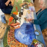 2 people are sat at a painting table, they are hiding brushes and painting