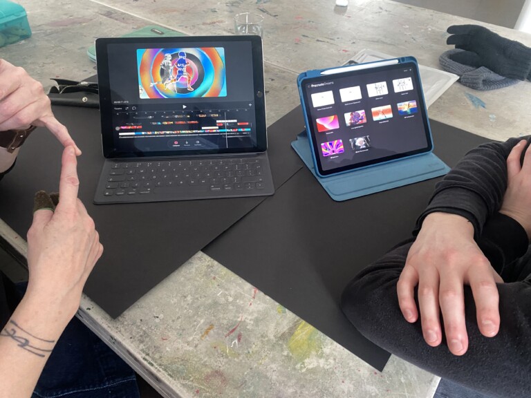 a close up of two iPads on a table. We can see two people, their hands are just visible in shot.