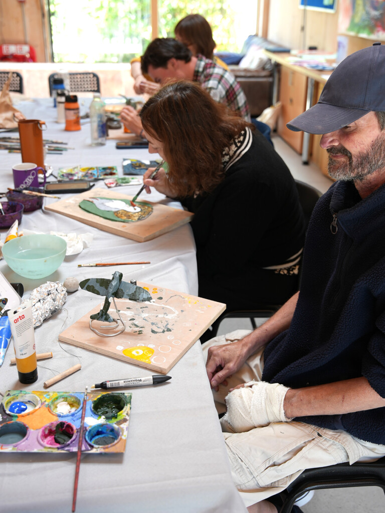 In a bright sunny room, a group of people sit around a table, they are working on small clay sculptures, the surface of the table is covered with art materials.