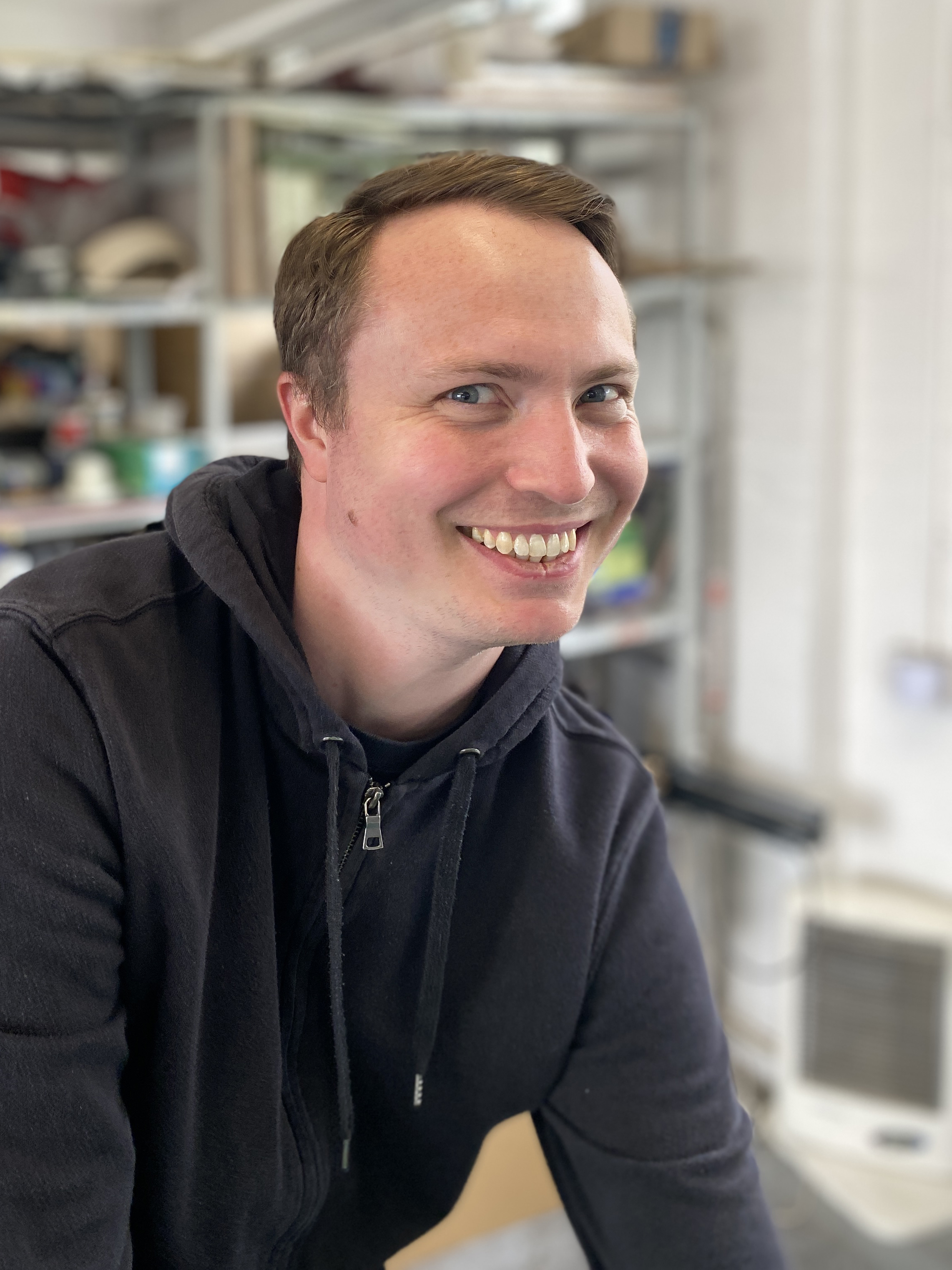 A man wearing a dark hoodie is looking into the camera and smiling